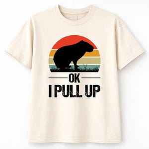 May include: Cream-colored t-shirt featuring a black capybara silhouette against a retro sunset design with orange, yellow, and teal stripes. The text "OK I PULL UP" is printed below the graphic in a bold, black font.
