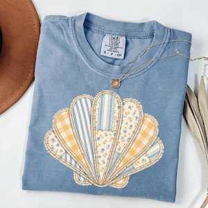 May include: A light blue Comfort Colors t-shirt featuring a seashell design. The seashell is made of various fabrics, including yellow gingham, blue stripes, and floral patterns. A gold necklace with a pendant rests on the shirt.