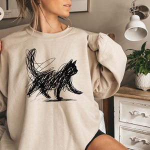 May include: Sand-colored sweatshirt featuring a black scribble-style cat design. The cat is depicted in a walking pose with a long tail. The sweatshirt has a crew neck and long sleeves. The word "SAND" is printed on a circular label in the upper left corner.