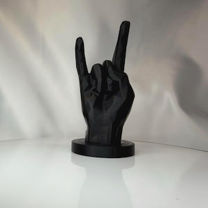 May include: A black, low-poly geometric sculpture of a hand making the rock and roll sign. The hand is positioned vertically with the index and pinky fingers extended upward, mounted on a circular black base with a smooth, matte finish.