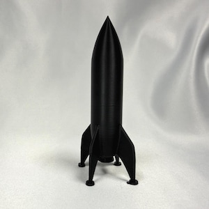 May include: A black rocket model with a pointed top and four stabilizing fins. The rocket stands upright on four small legs. The model is made of a solid, matte black material.