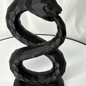 May include: A black, low-poly geometric sculpture of a coiled snake. The figure features a faceted, angular surface texture throughout its body, with the snake's head positioned at the top, curving downward to form a continuous, upright loop shape.