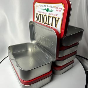 May include: A stack of rectangular metal Altoids tins with silver-colored bodies and red lids. The top lid features the white and red Altoids logo with the text Curiously Strong Mints and Peppermint. The tins are open, revealing empty interiors.