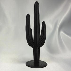 May include: A black, three-dimensional cactus sculpture with a textured surface. The cactus has a central, upright stem with three side arms, all rising from a circular base. The sculpture is a decorative item.