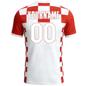 May include: A white and red football shirt featuring the text "YOURNAME" and the number "00". The shirt has a checkered design on the back and shoulders, with red sleeves and white trim.