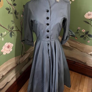May include: A vintage blue dress with a high collar and button-up front. The dress has a full skirt and three-quarter sleeves. It is on a mannequin against a floral wallpaper background.