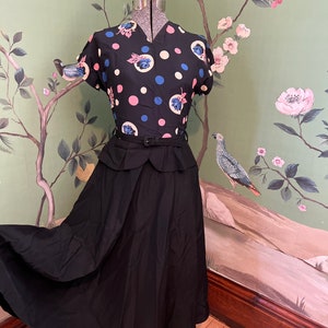 Könnte beinhalten: Ein schwarz-weißes Polka-Dot-Kleid mit Blumendruck. Das Kleid hat ein tailliertes Oberteil und einen vollen Rock. Das Kleid befindet sich auf einer Schaufensterpuppe vor einer grünen Blumen-Tapete.