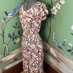 K&ouml;nnte beinhalten: Ein wei&szlig;es Kleid mit einem Blumendruck aus rosa Bl&uuml;ten und braunen Zweigen. Das Kleid hat einen runden Ausschnitt und kurze &Auml;rmel. Es ist an der Taille tailliert und flie&szlig;t zum Saum hin aus.
