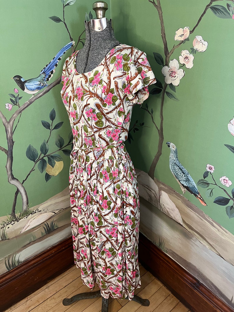 K&ouml;nnte beinhalten: Ein wei&szlig;es Kleid mit einem Blumendruck aus rosa Bl&uuml;ten und braunen Zweigen. Das Kleid hat einen runden Ausschnitt und kurze &Auml;rmel. Es ist an der Taille tailliert und flie&szlig;t zum Saum hin aus.