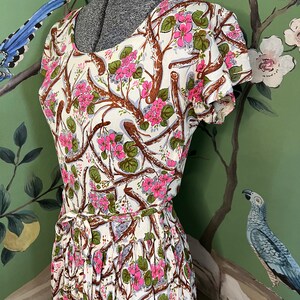 K&ouml;nnte beinhalten: Ein wei&szlig;es Kleid mit einem Blumendruck aus rosa Bl&uuml;ten und braunen Zweigen. Das Kleid hat kurze &Auml;rmel und einen taillierten Oberteil. Der Rock ist weit und an der Taille gerafft.