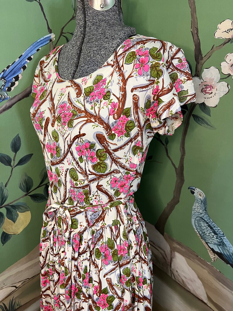 K&ouml;nnte beinhalten: Ein wei&szlig;es Kleid mit einem Blumendruck aus rosa Bl&uuml;ten und braunen Zweigen. Das Kleid hat kurze &Auml;rmel und einen taillierten Oberteil. Der Rock ist weit und an der Taille gerafft.