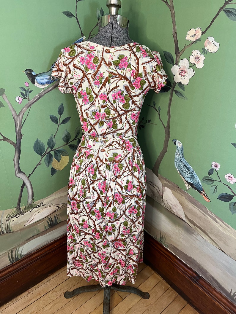 K&ouml;nnte beinhalten: Ein wei&szlig;es Kleid mit einem Blumendruck aus rosa Bl&uuml;ten und braunen Zweigen. Das Kleid hat kurze &Auml;rmel und ein tailliertes Oberteil. Das Kleid befindet sich auf einer Schaufensterpuppe vor einem gr&uuml;nen Hintergrund mit einem floralen Tapetenmuster.