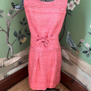 Puede incluir: Vestido rosa coral sin mangas con tejido texturizado. El vestido presenta un corpiño ajustado, un lazo en la cintura y una falda a la rodilla. El vestido se exhibe en un maniquí.