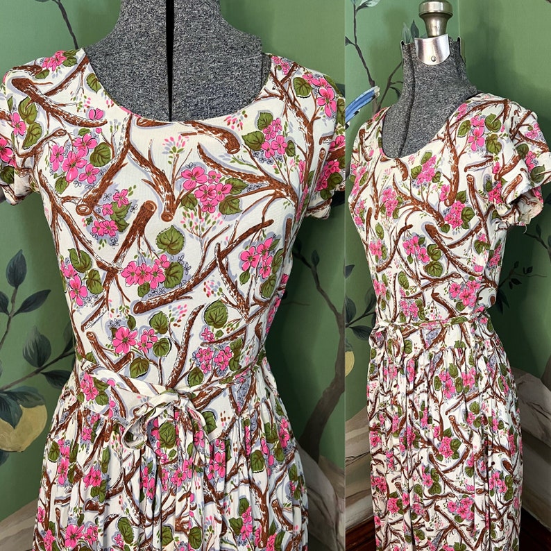 K&ouml;nnte beinhalten: Ein wei&szlig;es Kleid mit rosafarbenem Blumendruck mit &Auml;sten und Bl&auml;ttern. Das Kleid hat einen runden Ausschnitt, kurze &Auml;rmel und eine Schleife in der Taille.