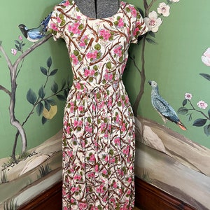 K&ouml;nnte beinhalten: Ein wei&szlig;es Midikleid mit einem Blumendruck mit rosa Bl&uuml;ten, braunen Zweigen und gr&uuml;nen Bl&auml;ttern. Das Kleid hat einen runden Ausschnitt, kurze &Auml;rmel und einen gerafften Rock.