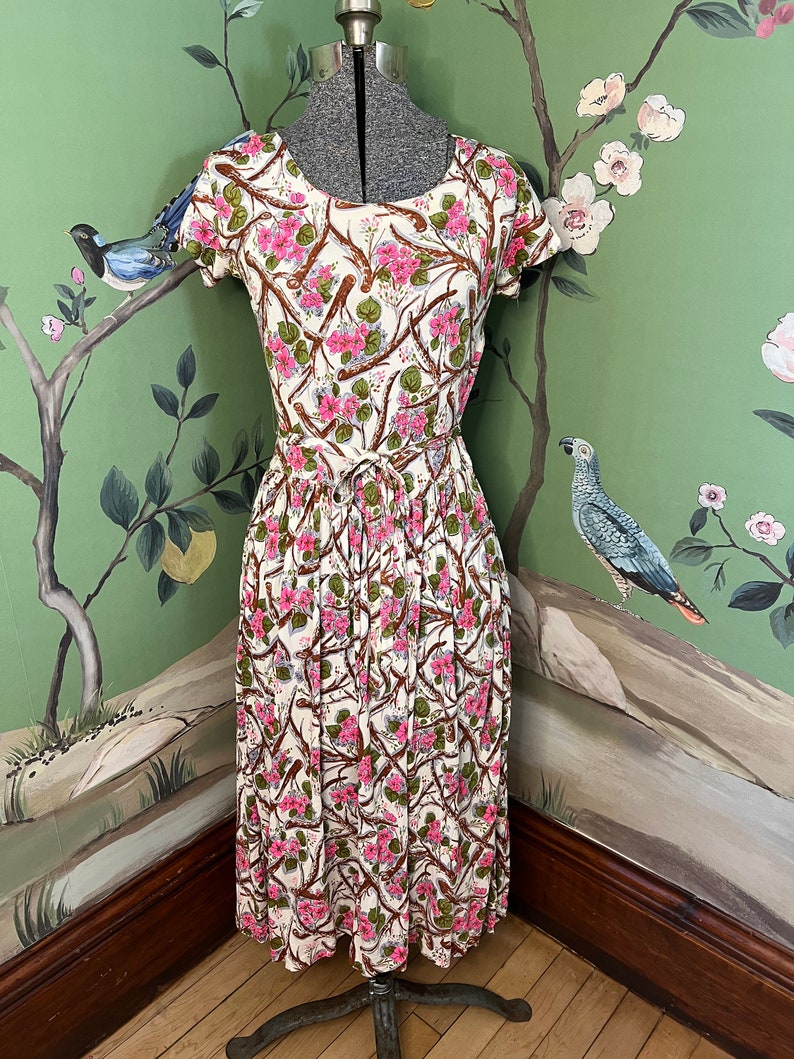 K&ouml;nnte beinhalten: Ein wei&szlig;es Midikleid mit einem Blumendruck mit rosa Bl&uuml;ten, braunen Zweigen und gr&uuml;nen Bl&auml;ttern. Das Kleid hat einen runden Ausschnitt, kurze &Auml;rmel und einen gerafften Rock.