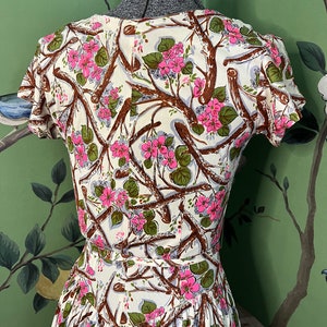 K&ouml;nnte beinhalten: Ein wei&szlig;es Kleid mit einem Blumendruck aus rosa Bl&uuml;ten und braunen Zweigen. Das Kleid hat kurze &Auml;rmel und einen taillierten Oberteil.