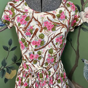 K&ouml;nnte beinhalten: Ein wei&szlig;es Kleid mit einem Blumendruck aus rosa Bl&uuml;ten und braunen Zweigen. Das Kleid hat einen runden Ausschnitt und kurze &Auml;rmel. Das Kleid ist in der Taille mit einer passenden Schleife gebunden.