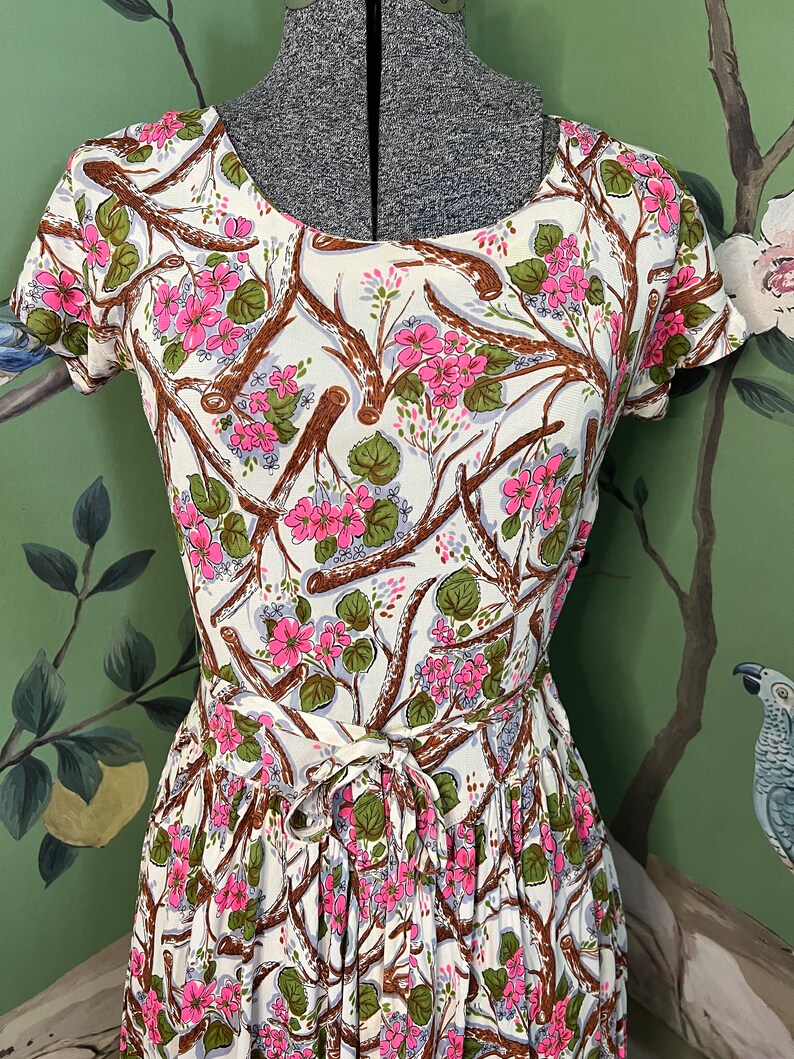 K&ouml;nnte beinhalten: Ein wei&szlig;es Kleid mit einem Blumendruck aus rosa Bl&uuml;ten und braunen Zweigen. Das Kleid hat einen runden Ausschnitt und kurze &Auml;rmel. Das Kleid ist in der Taille mit einer passenden Schleife gebunden.