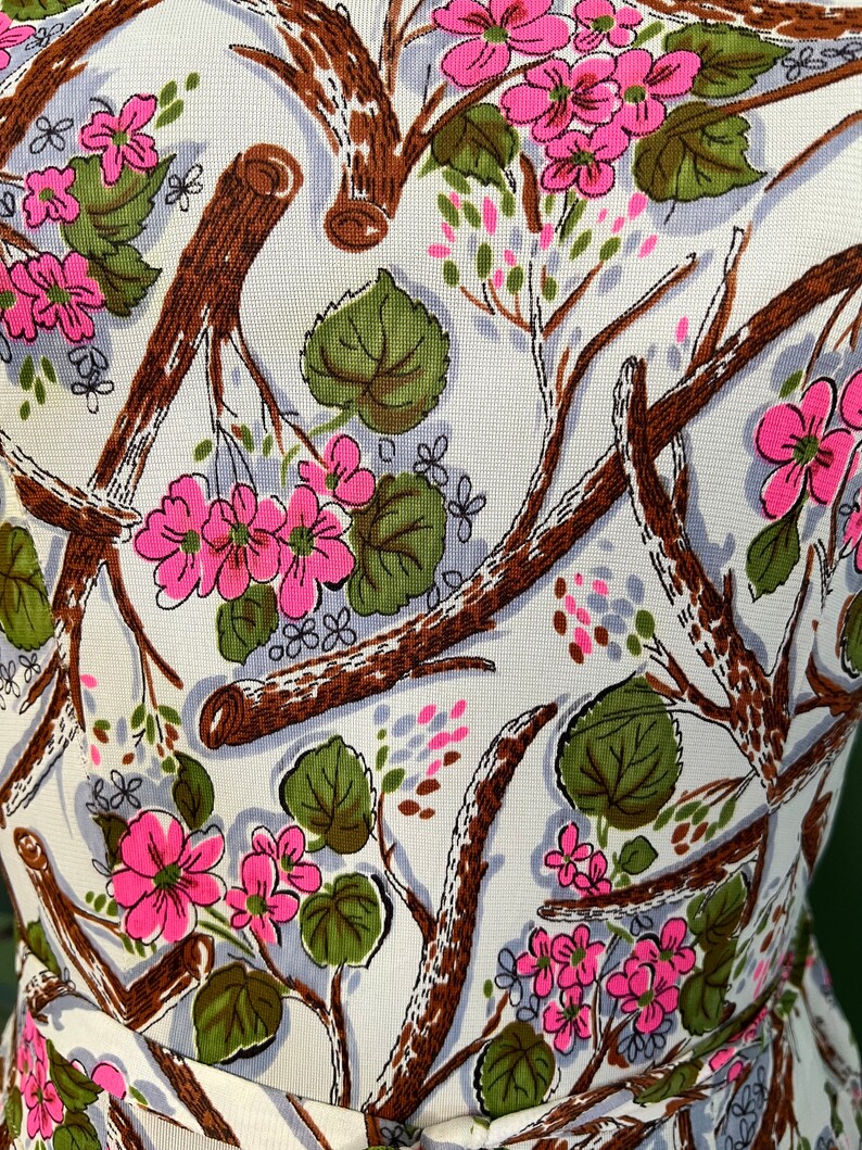 K&ouml;nnte beinhalten: Ein wei&szlig;er Stoff mit einem sich wiederholenden Muster aus rosa Bl&uuml;ten, braunen Zweigen und gr&uuml;nen Bl&auml;ttern.