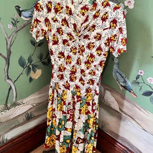 Könnte beinhalten: Ein weißes Kleid mit einem Blumendruck mit roten, gelben und grünen Blumen. Das Kleid hat einen V-Ausschnitt und kurze Ärmel. Das Kleid ist am Rock gefaltet.