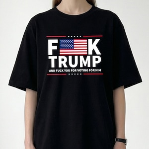 Puede incluir: Camiseta negra con un diseño gráfico que presenta el texto "F**K TRUMP" en letras mayúsculas blancas, con una bandera estadounidense. El texto "AND F**K YOU FOR VOTING FOR HIM" está debajo.