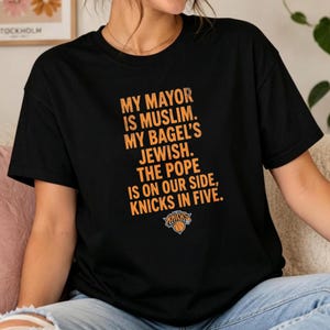 Op de afbeelding: Een zwart T-shirt met korte mouwen en oranje tekst: MY MAYOR IS MUSLIM. MY BAGEL'S JEWISH. THE POPE IS ON OUR SIDE, KNICKS IN FIVE. Onder de tekst staat een klein oranje en blauw logo van het basketbalteam de New York Knicks.