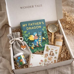 Puede incluir: Una caja de regalo con el texto "WONDER TALE" en la tapa, que contiene un libro titulado "MY FATHER'S DRAGON", lápices de colores, pegatinas, una lupa, una pequeña bolsa con un dije de zorro y un tarro de caramelos en forma de estrella. La caja está llena de papel triturado.