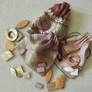 May include: Two small fabric pouches with drawstring closures, filled with various wooden and shell-like shapes. The pouches are decorated with gold embroidery and have a brown and beige color scheme.