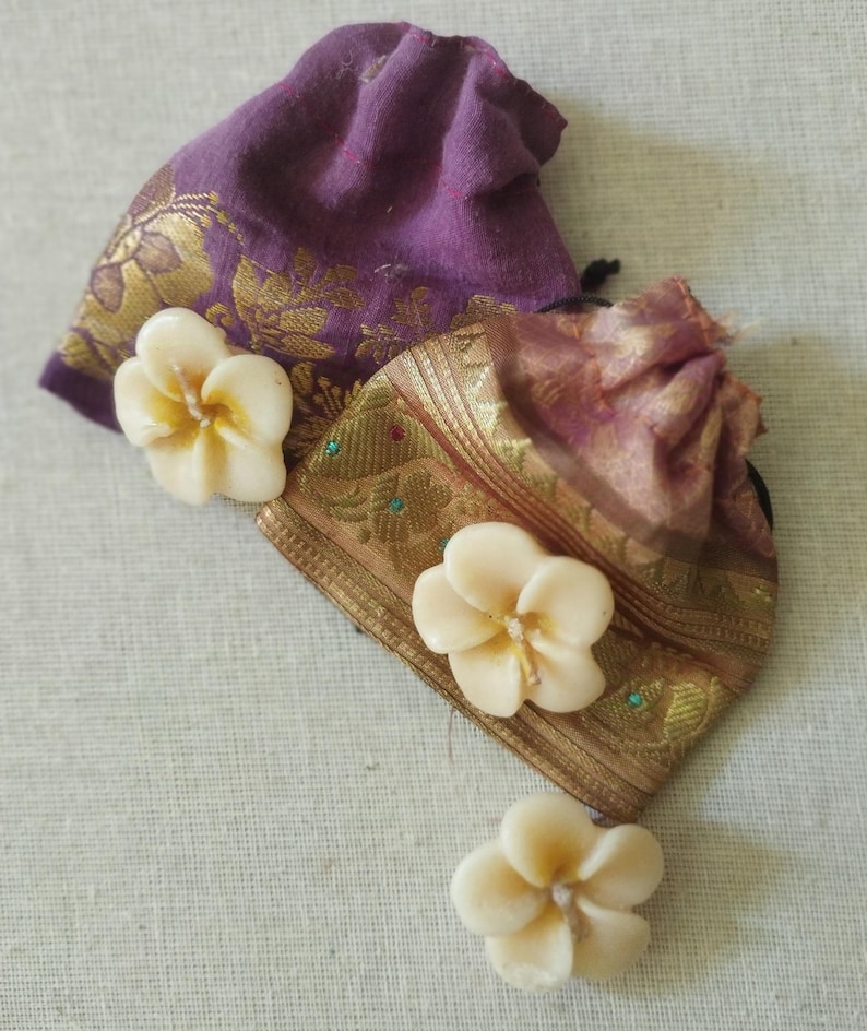 May include: Three white flower-shaped candles with a gold and purple fabric pouch in the background.