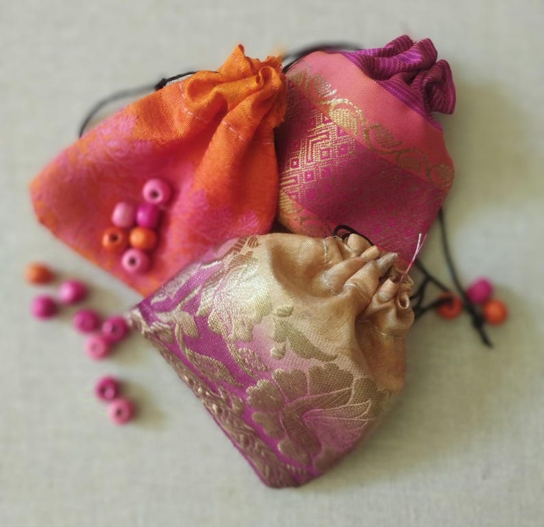 May include: Three small drawstring bags made of colorful patterned fabric. The bags are pink, orange, and gold with intricate designs. The bags are filled with pink wooden beads.