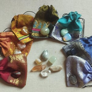 May include: Ten small pouches made of colorful fabric with drawstring closures, each containing a different gemstone. The pouches are arranged in a circle on a beige surface.