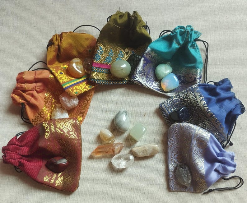 May include: Ten small pouches made of colorful fabric with drawstring closures, each containing a different gemstone. The pouches are arranged in a circle on a beige surface.