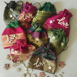 May include: A collection of eight small, colorful drawstring bags made of fabric with gold accents. Each bag has a wooden cutout of the word "love" attached to the front. The bags are surrounded by small wooden stars.