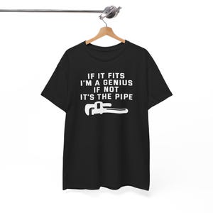 Puede incluir: Camiseta negra con el texto blanco "IF IT FITS I'M A GENIUS IF NOT IT'S THE PIPE" y un dibujo blanco de una llave inglesa. La camiseta está colgada de una percha de madera.