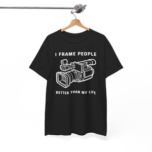 May include: Black t-shirt featuring a white video camera graphic and the text "I FRAME PEOPLE BETTER THAN MY LIFE". The garment is suspended from a wooden hanger.