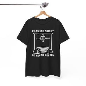 May include: Black t-shirt with a white graphic of a 3D printer and the text "FILAMENT ADDICT" above and "NO REHAB NEEDED" below. The shirt is hanging on a wooden hanger.