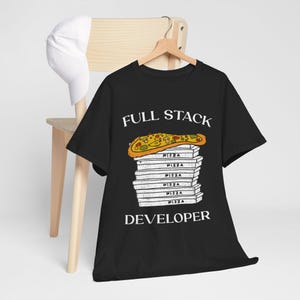 Op de afbeelding: Zwart t-shirt met de tekst "FULL STACK DEVELOPER" en een pizzapunt-afbeelding. De pizzapunt ligt bovenop een stapel pizzadozen. Een witte baseballpet hangt aan een houten stoel.