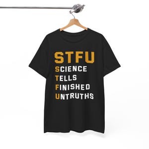 Puede incluir: Camiseta negra con el acrónimo "STFU" en letras doradas. Debajo del acrónimo, las palabras "SCIENCE TELLS FINISHED UNTRUTHS" están impresas en blanco. La camiseta está colgada en una percha de madera.