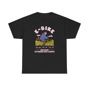 Puede incluir: Camiseta negra con un gráfico de bicicleta eléctrica. El diseño incluye el texto "E-BIKE" y "BECAUSE - PATIENCE HAS LIMITS". La ilustración muestra a una persona con una chaqueta azul en una bicicleta amarilla y verde. El diseño es de estilo caricatura.