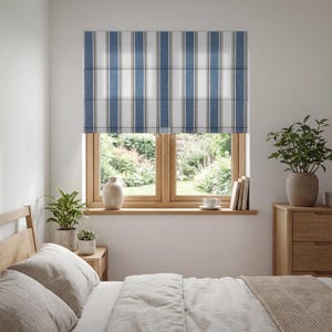 May include: A bedroom window with a blue and white striped Roman shade. The window is framed by a wooden frame, and a wooden nightstand with plants and a vase is visible. The bed has white bedding.