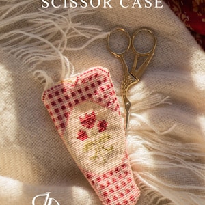 Puede incluir: Un estuche para tijeras de punto de cruz con un patrón de cuadros rojos y blancos y bordados florales. Unas tijeras doradas descansan junto al estuche sobre una tela suave, blanca y con flecos. El texto "Cross Stitch Pattern SCISSOR CASE" es visible.