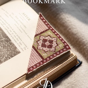 Può includere: Un segnalibro fatto a mano a punto croce con un design geometrico nei toni del rosa, verde e bordeaux. Il segnalibro è triangolare e posto in un libro aperto. Il testo "Cross Stitch Pattern BOOKMARK" è visibile.