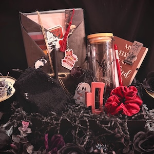 Puede incluir: Una caja de regalo de temática oscura llena de varios artículos. Incluye un libro, pegatinas, un bolígrafo, una pinza para el pelo, una goma para el pelo, un tarro y flores decorativas. La paleta de colores es principalmente negra, roja y blanca.
