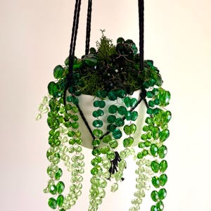 May include: A white hanging planter containing a decorative plant made of faceted green glass beads on thin wires. The beads transition from dark emerald to light lime green, mimicking trailing vines. The planter is suspended by black cords.