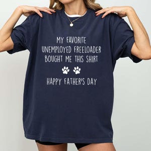 Könnte beinhalten: Marineblaues T-Shirt mit weißem Text: "MY FAVORITE UNEMPLOYED FREELOADER BOUGHT ME THIS SHIRT" und "HAPPY FATHER'S DAY" mit Pfotenabdrücken. Das T-Shirt hat einen lässigen Stil.