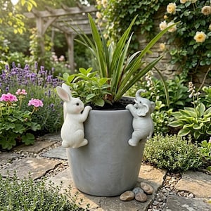 May include: A grey ceramic plant pot with a rabbit and elephant figurine attached. The pot contains green plants and is placed on a stone path. The background shows a garden with flowers and a wooden structure.
