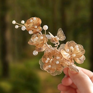 May include: A gold-toned hair accessory featuring multiple wire-formed flowers and a small butterfly. The piece is embellished with numerous small, round, white faux pearls attached to thin wire stems, creating a delicate, layered floral arrangement.