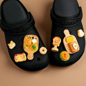 May include: Black clogs decorated with colorful food-themed charms, including miniature burgers, donuts, and toast. The charms are attached to the top of the shoes, adding a playful and unique touch to the footwear. The clogs are made of a durable material.