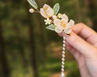 May include: A hair clip featuring translucent pale pink flower petals, light green leaves, and small gold-toned metal flowers. The design includes a dangling strand of small white pearls and a single pearl accent on a thin gold wire stem.